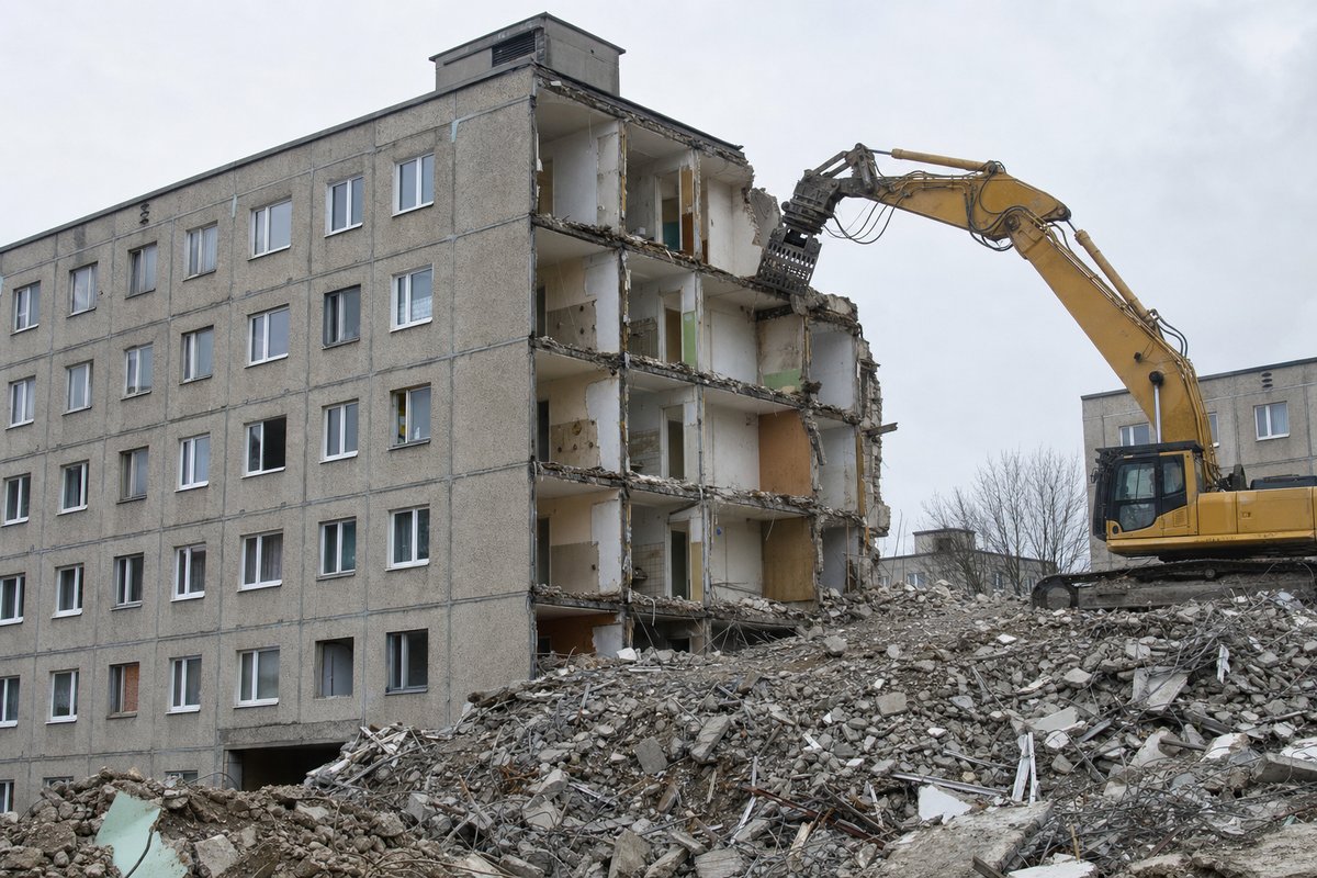 Teilweise abgerissener Plattenbau, ein Wandsegment entfernt und Innenräume sichtbar, Bagger mit Abbruchgreifer, flaches sächsisches Licht.